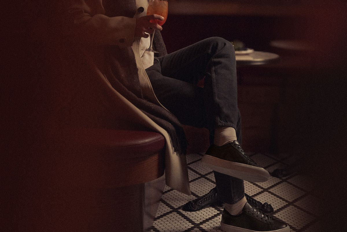 Man seated in a dimly lit lounge wearing Capri black low-top sneakers and holding a drink