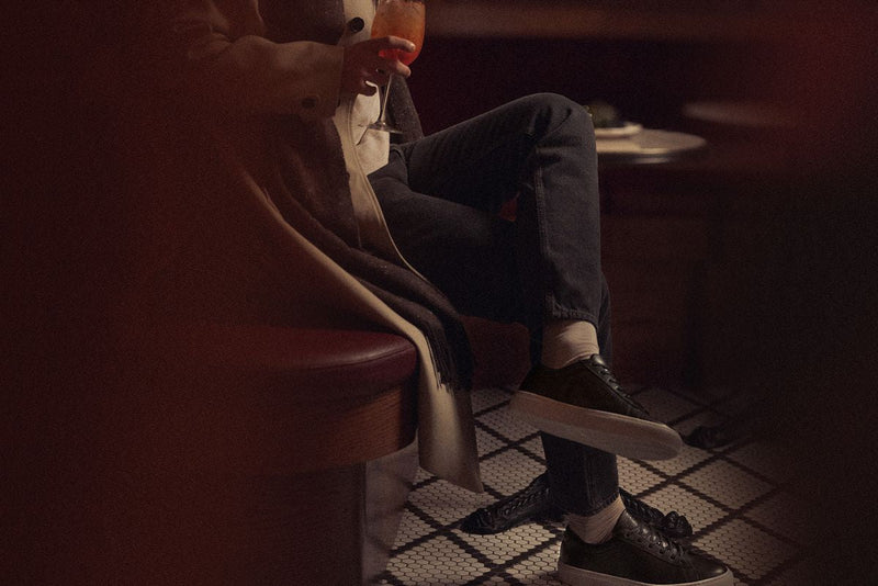 Man seated in a dimly lit lounge wearing Capri black low-top sneakers and holding a drink