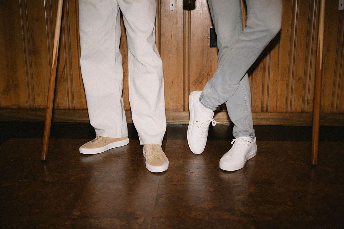 Two people wearing beige and white Capri sneakers stand on a polished wood floor in a wood-paneled room, holding pool cues