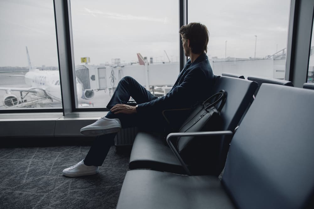Man wearing the Capri Triple White low-top sneakers sits in an airport lounge beside a window overlooking an airplane