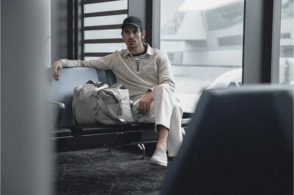 Man seated in a neutral smart-casual outfit with a duffel bag in a modern lounge, wearing Koio slip-on sneakers