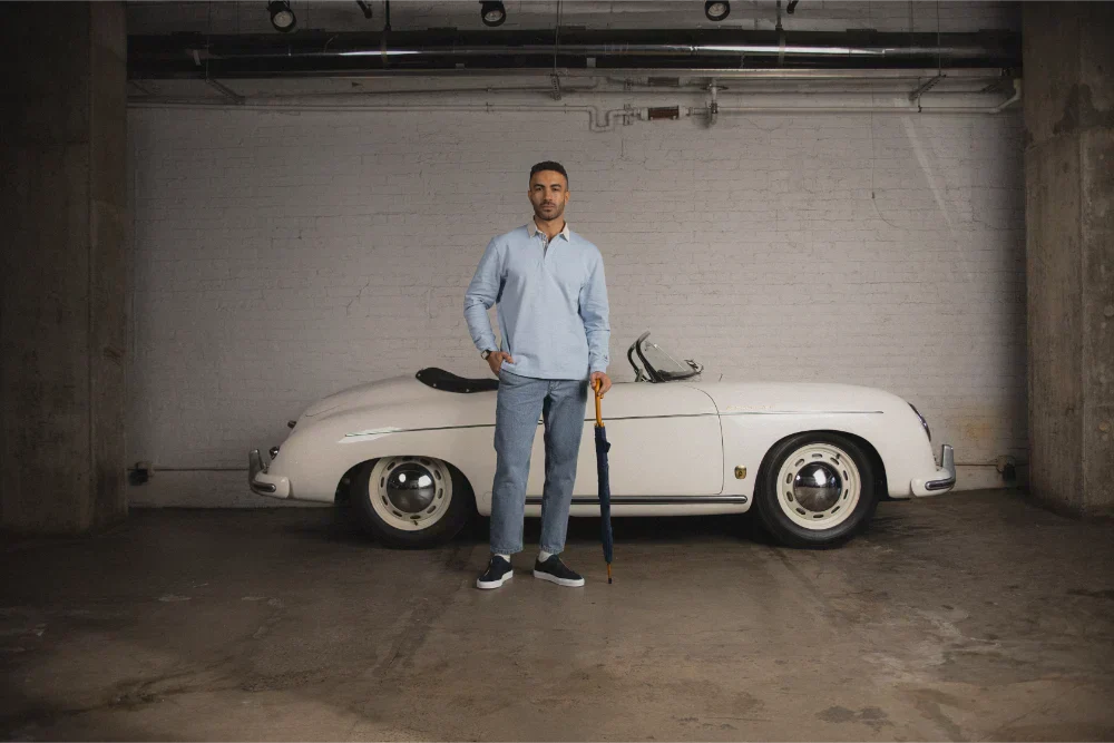 Man wearing Capri black low-top sneakers and jeans in front of a vintage car in a garage, holding an umbrella