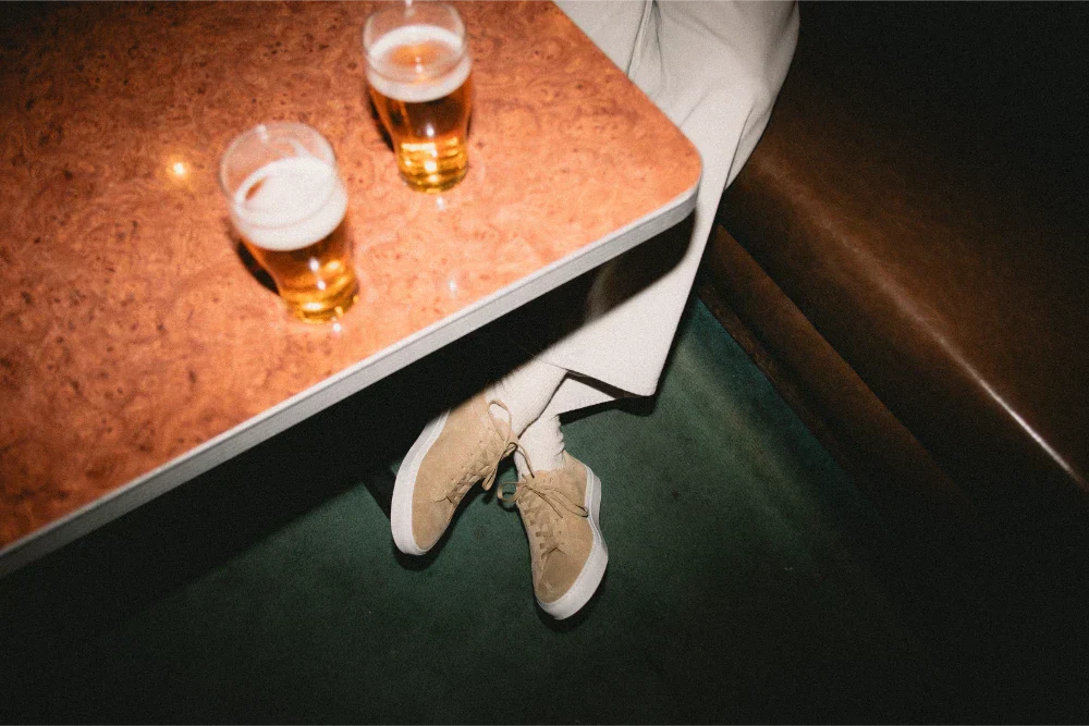 Person wearing Capri suede beige sneakers and white socks sits beside a marble table with two beers