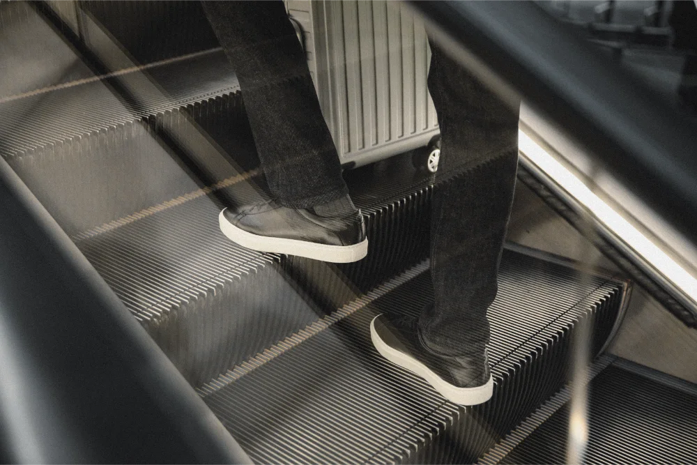 Traveler wearing black Capri Koio low-top sneakers rides an escalator