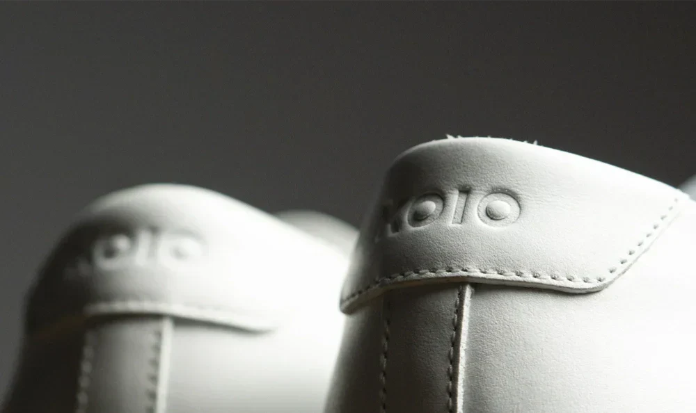 Close-up of Koio Capri Triple White low-top sneakers showing embossed logos and detailed stitching against a dark background
