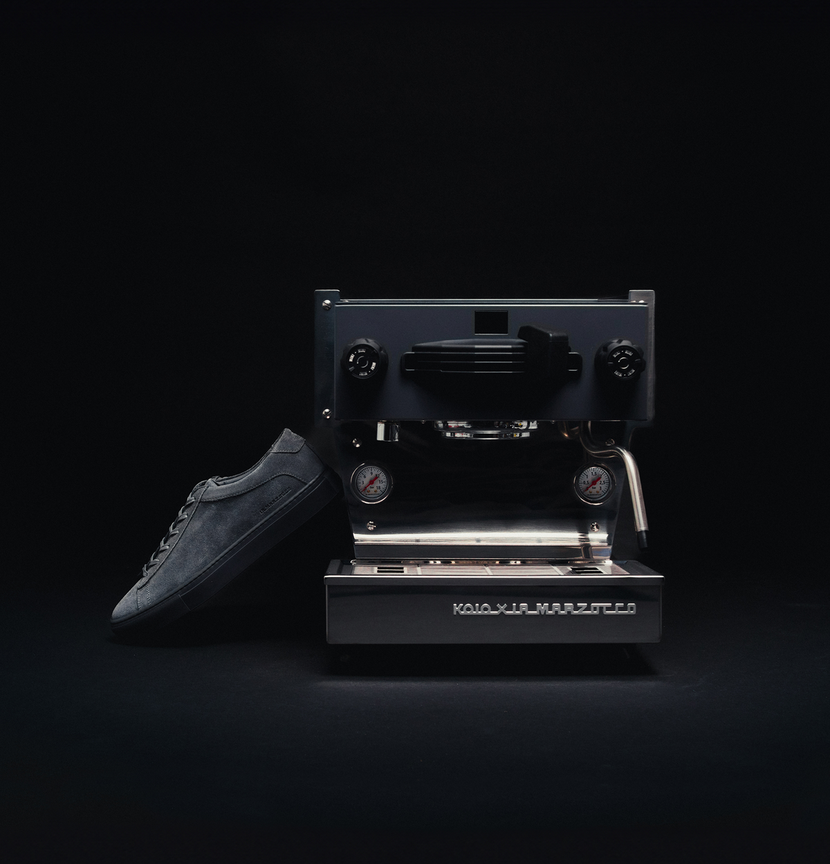 Grey suede low-top sneaker beside a La Marzocco espresso machine on a black background