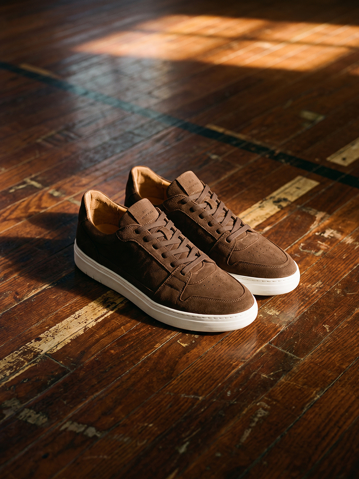 A pair of men's brown European suede low-top lace-up sneakers with white rubber soles, displayed on a rustic wooden floor with warm sunlight casting shadows.