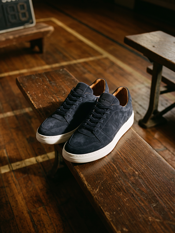 A pair of men's dark navy blue European suede lace-up sneakers with white rubber soles, resting on a rustic wooden bench in a warm, dimly lit room with wooden floors.