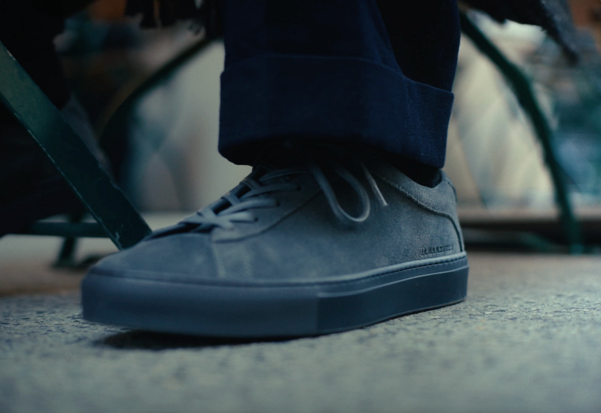 Grey suede low-top sneaker worn on pavement in an urban setting