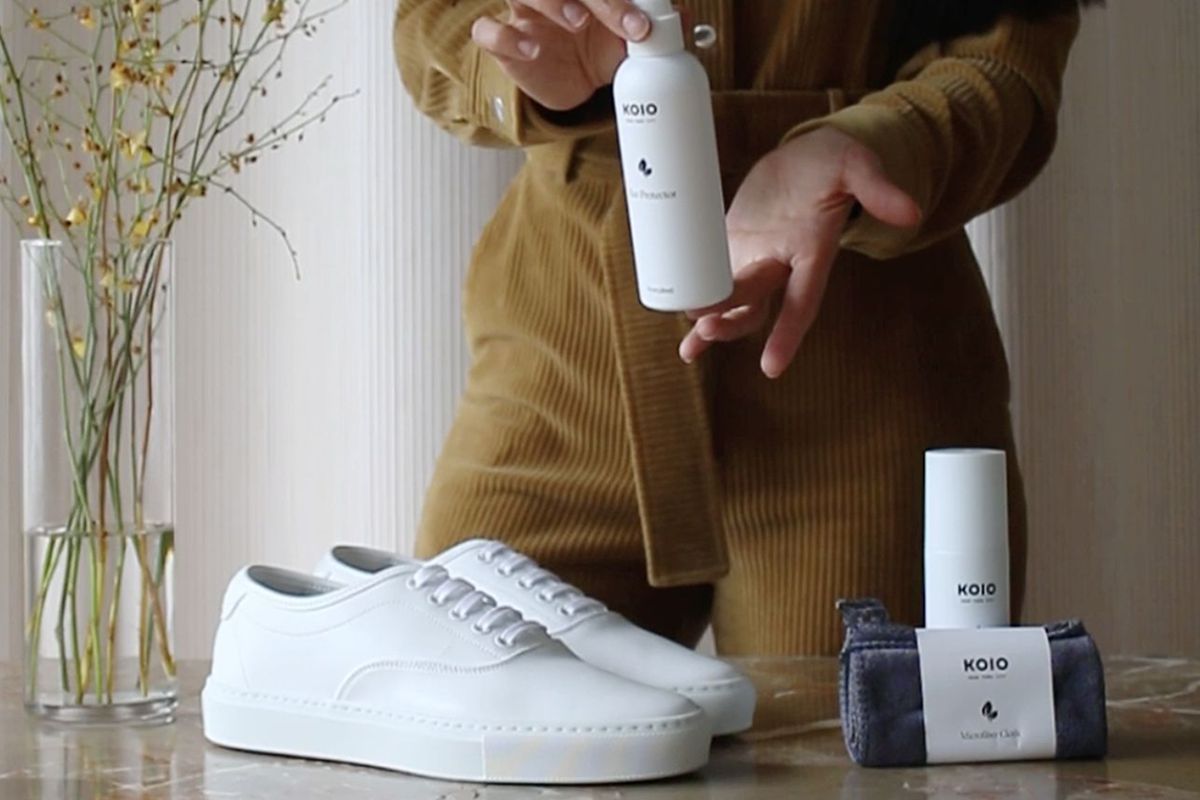 Person holding shoe cleaner above the Portofino white low-top sneakers and other Koio shoe care products on a table