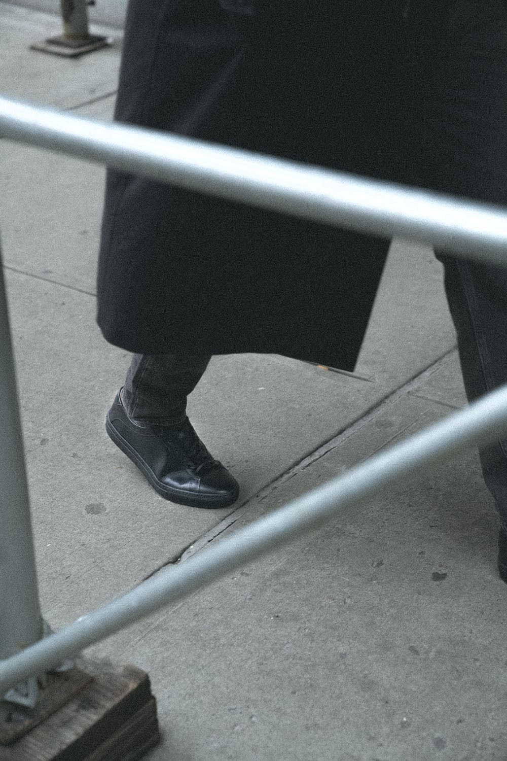 Person wearing a black leather sneaker with a tonal black outsole on a city sidewalk, viewed through a railing
