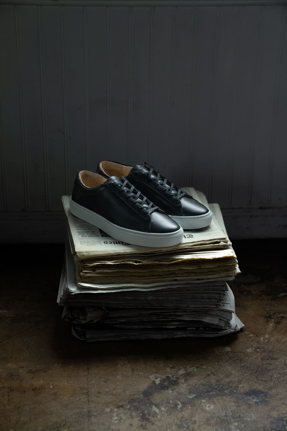Three-quarter view of black low-top sneakers showcasing white rubber soles on stacked newspapers