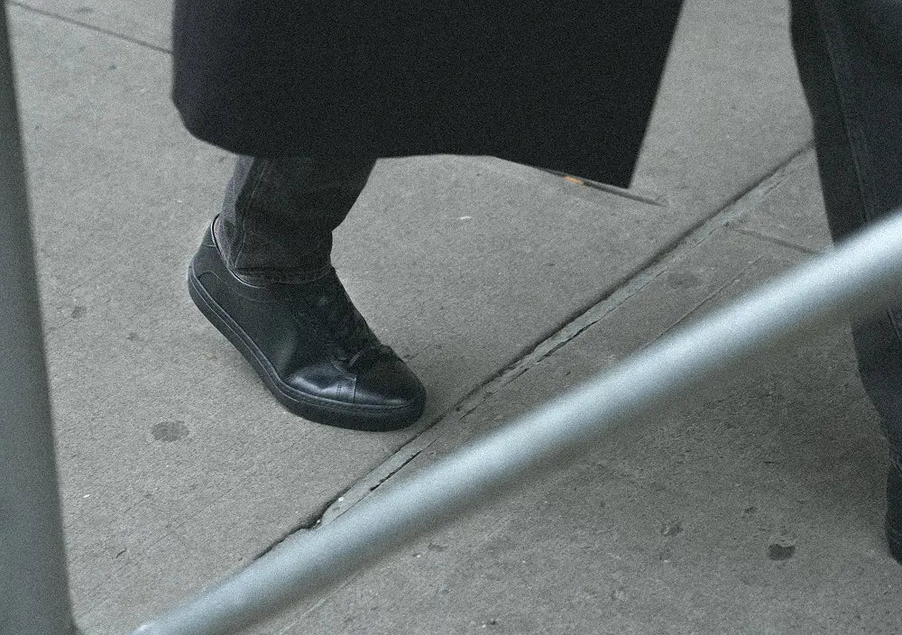 Person wearing Capri black leather sneakers walking on a city sidewalk, shown through scaffolding 