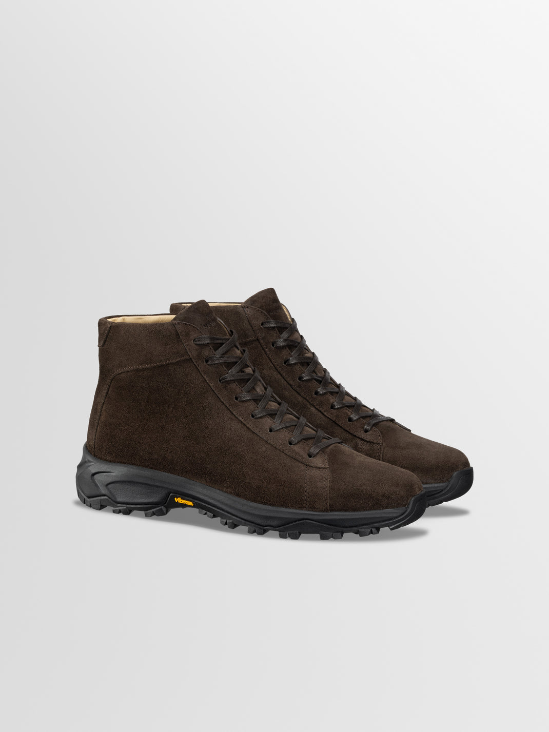 Three-quarter view of Aosta in Cacao boots showcasing brown suede uppers, black Vibram lug soles, and laces against a light gray background