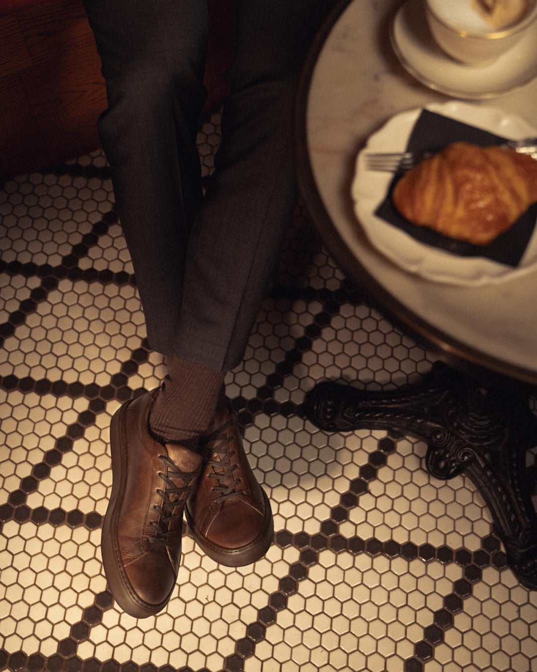Overhead view of Capri Antica brown leather low-top sneakers with tonal laces worn with dark trousers on patterned tile flooring