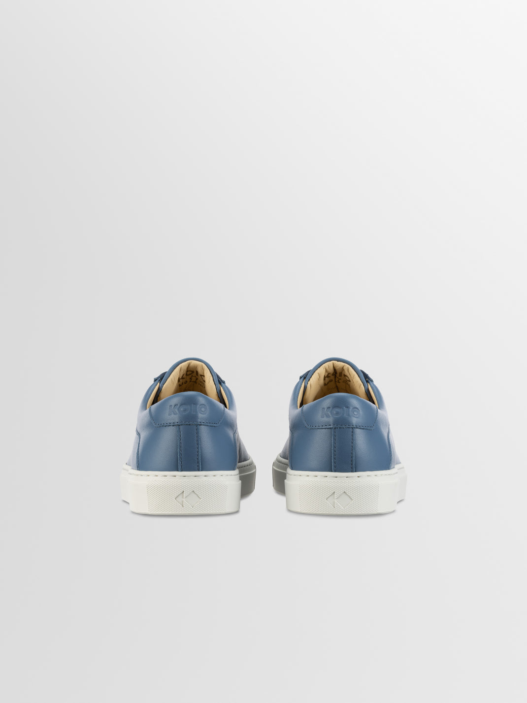 Rear view of Capri in Bay men’s blue leather low-top sneakers showcasing white rubber soles against a light gray background