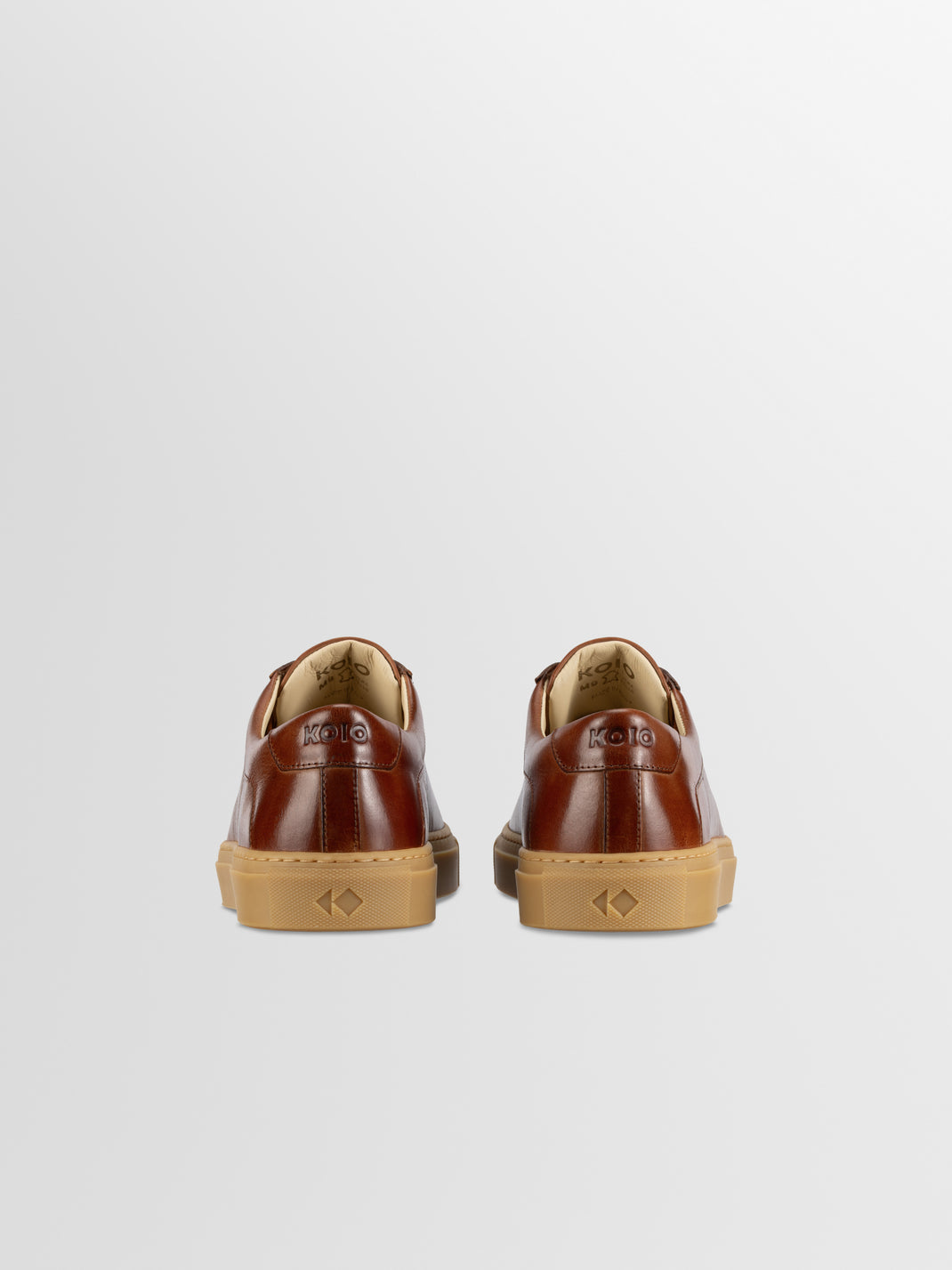 Rear view of brown leather Capri low-top sneakers showing KOIO heel logos and gum rubber soles on a light gray background