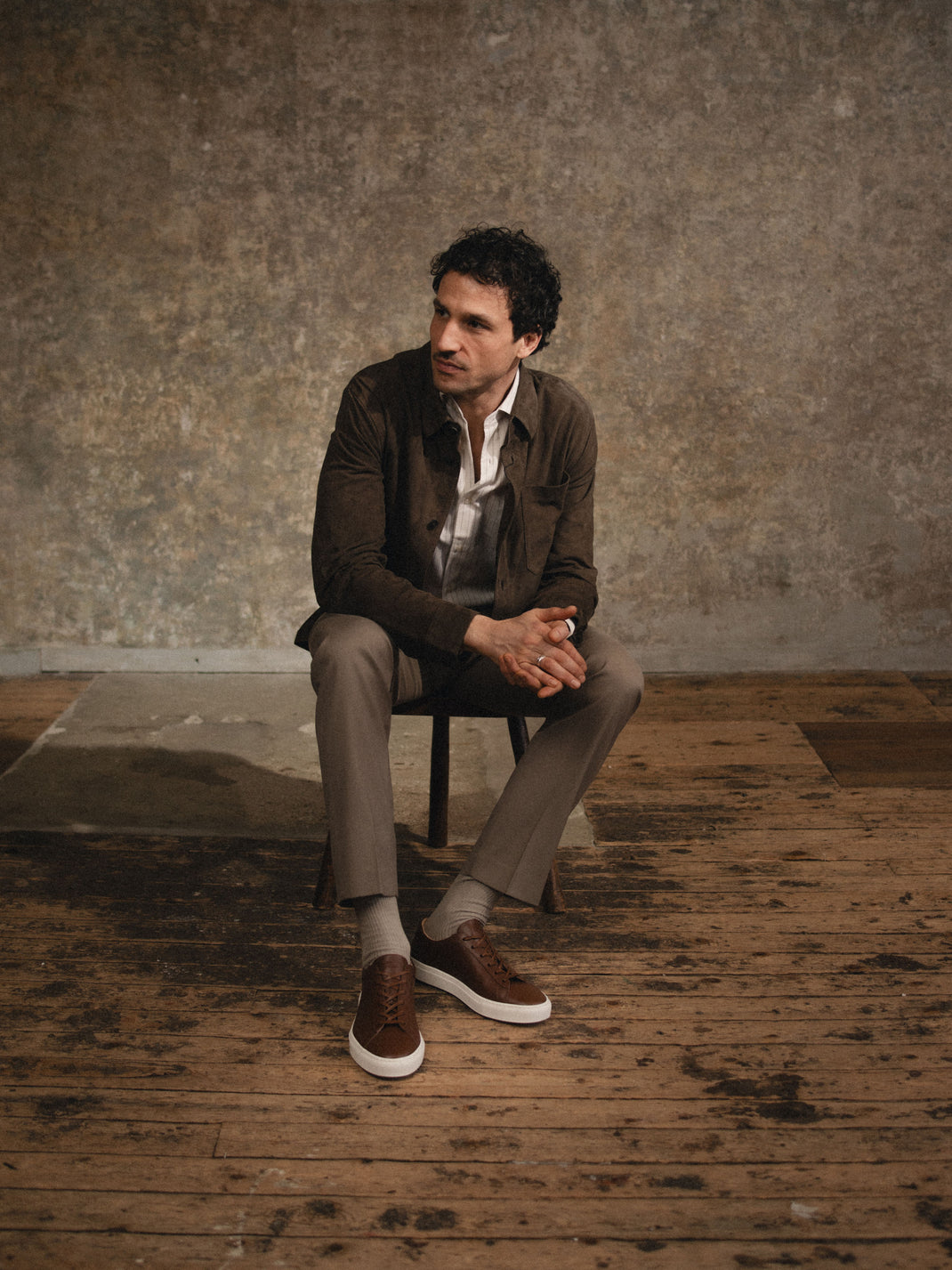 Man sitting on a chair with a white button down shirt, brown jacket, brown trousers, and brown leather sneakers with matching laces and white rubber soles