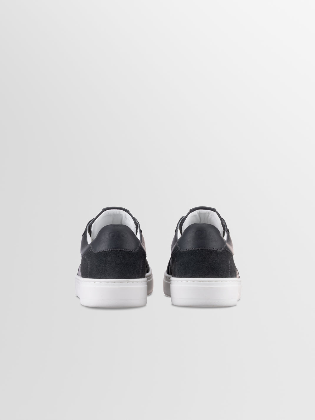 Rear view of Carrara in Basalt men’s black suede sneaker showcasing white rubber soles on a light gray background