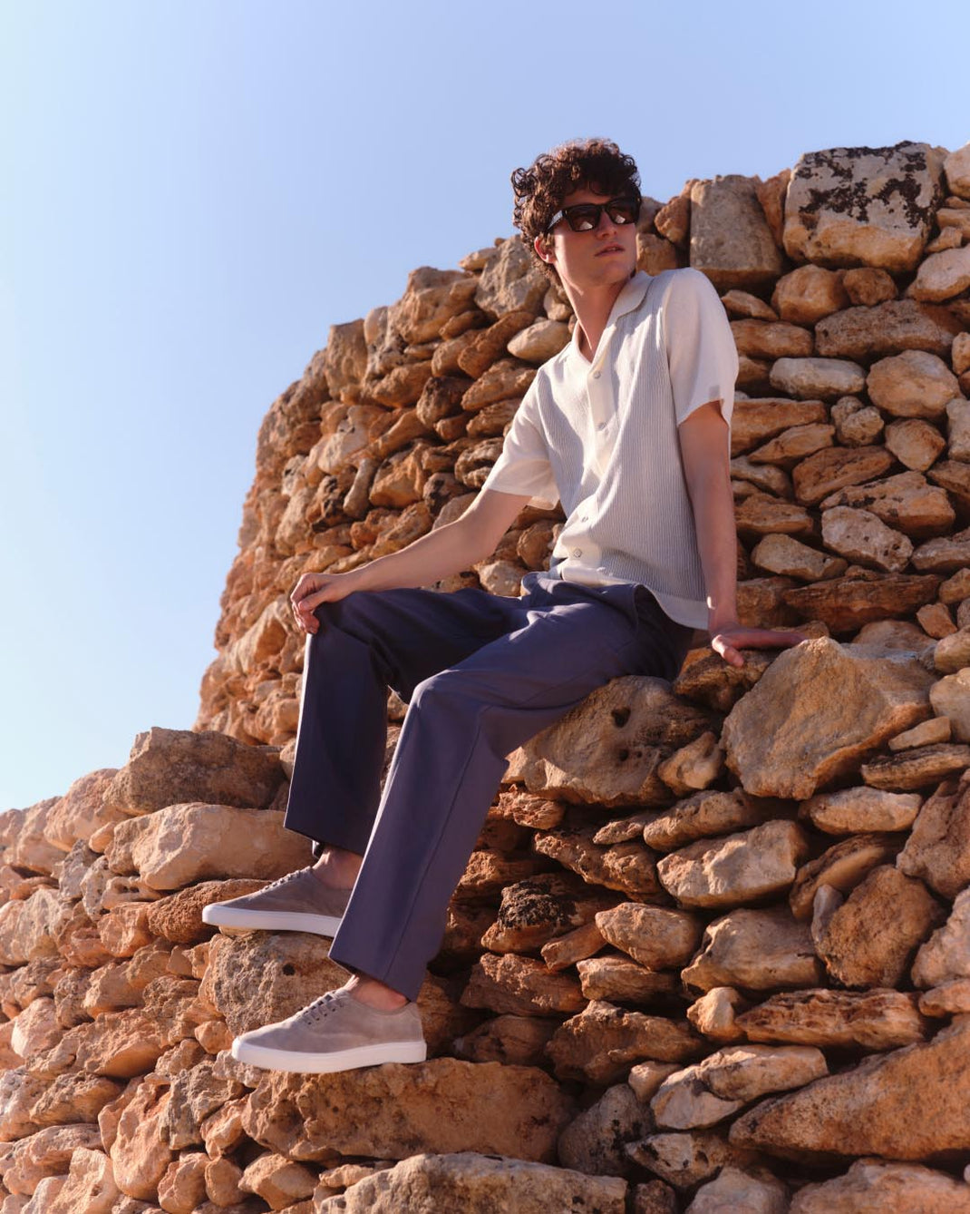 Man wearing beige grey Portofino suede sneakers while seated on sunlit rocks
