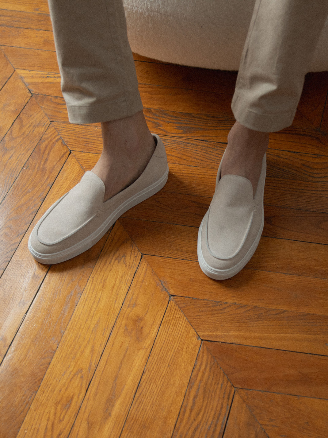Angled view of men’s beige suede Lido in Ivory slip-on loafers worn with beige trousers on a parquet floor