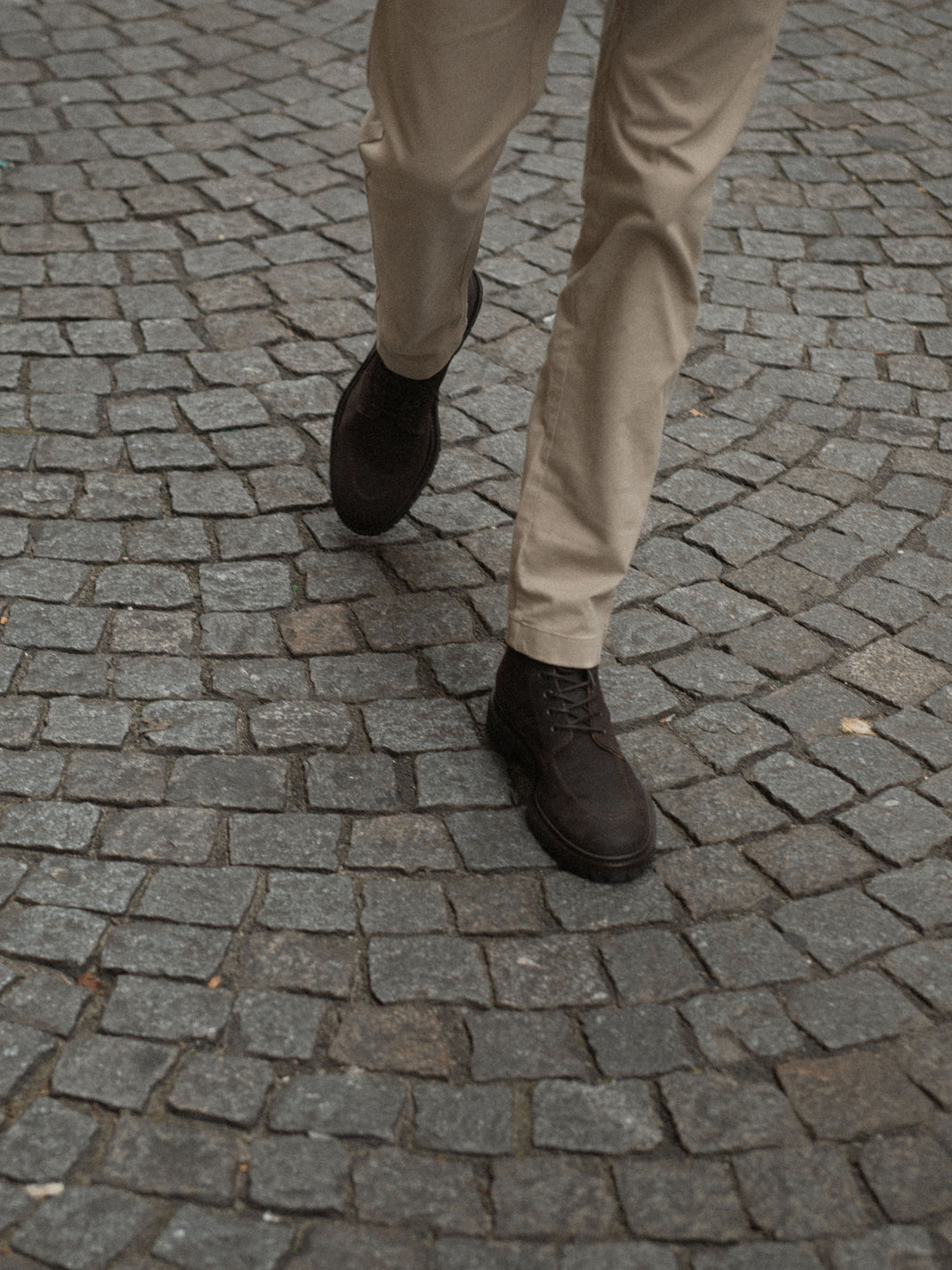 Person wearing dark brown Milo suede boots with beige trousers on a cobblestone street