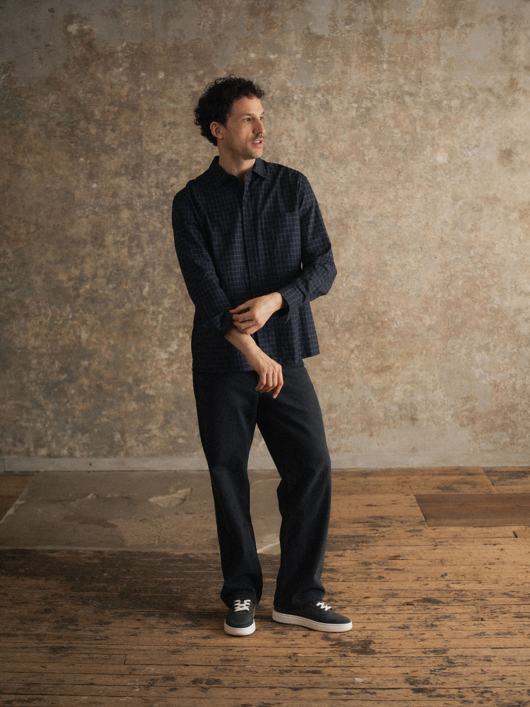 Man standing in black suede Carrara sneakers on a wooden floor against a textured wall