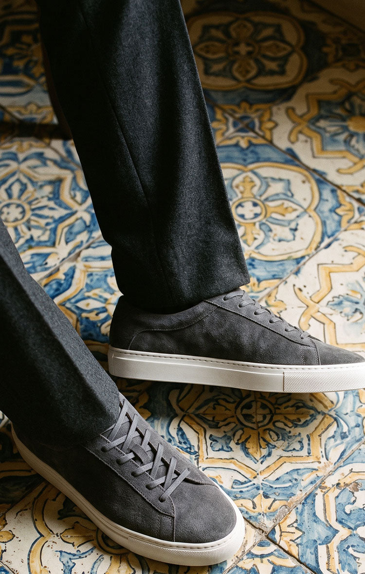 Close-up of Capri in Roccia men’s grey suede low-top sneakers showcasing white rubber soles with patterned tile background