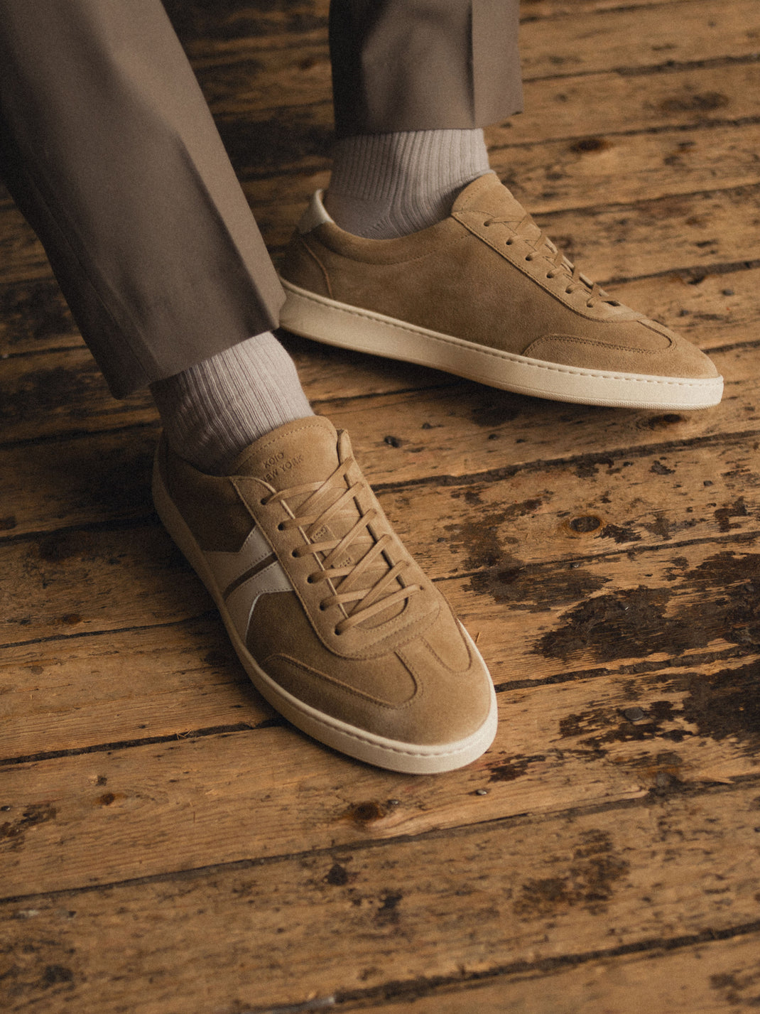 Close-up view of Monza in Wheat light brown suede low-top sneakers with lace-up fronts, worn on a wooden floor