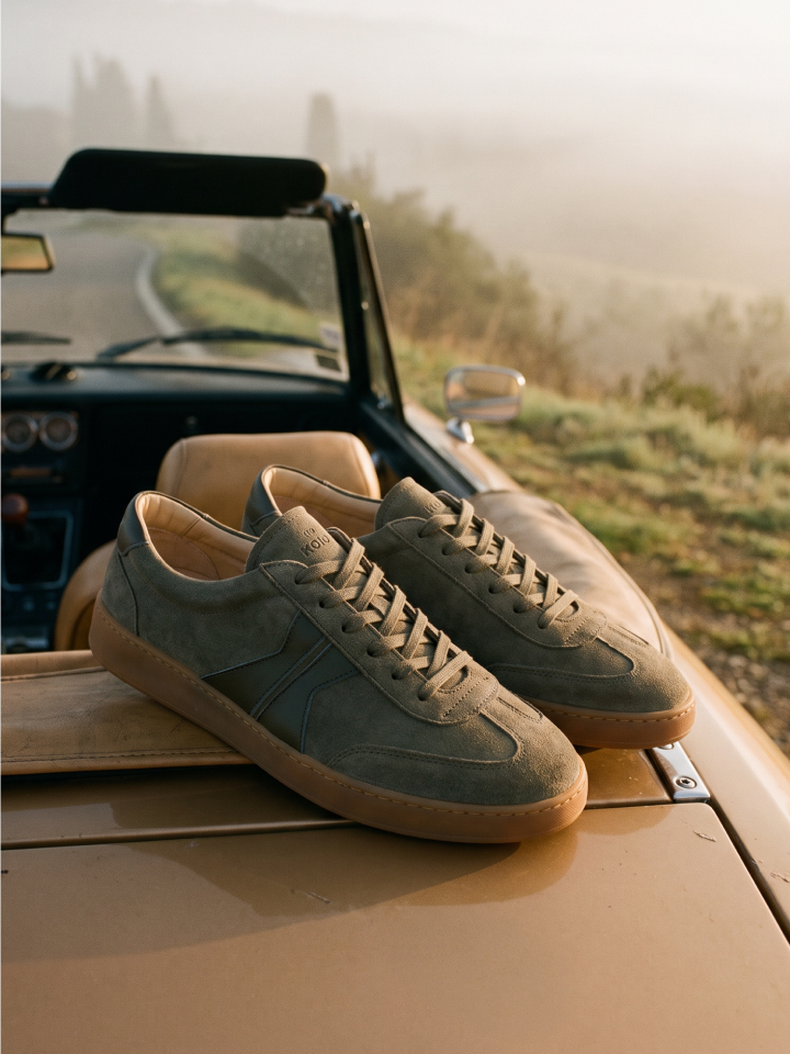 A pair of men's Italian leather suede muted gray-green lace-up low-top sneakers resting on the hood of a vintage tan convertible car, with a misty country road and trees in the background.