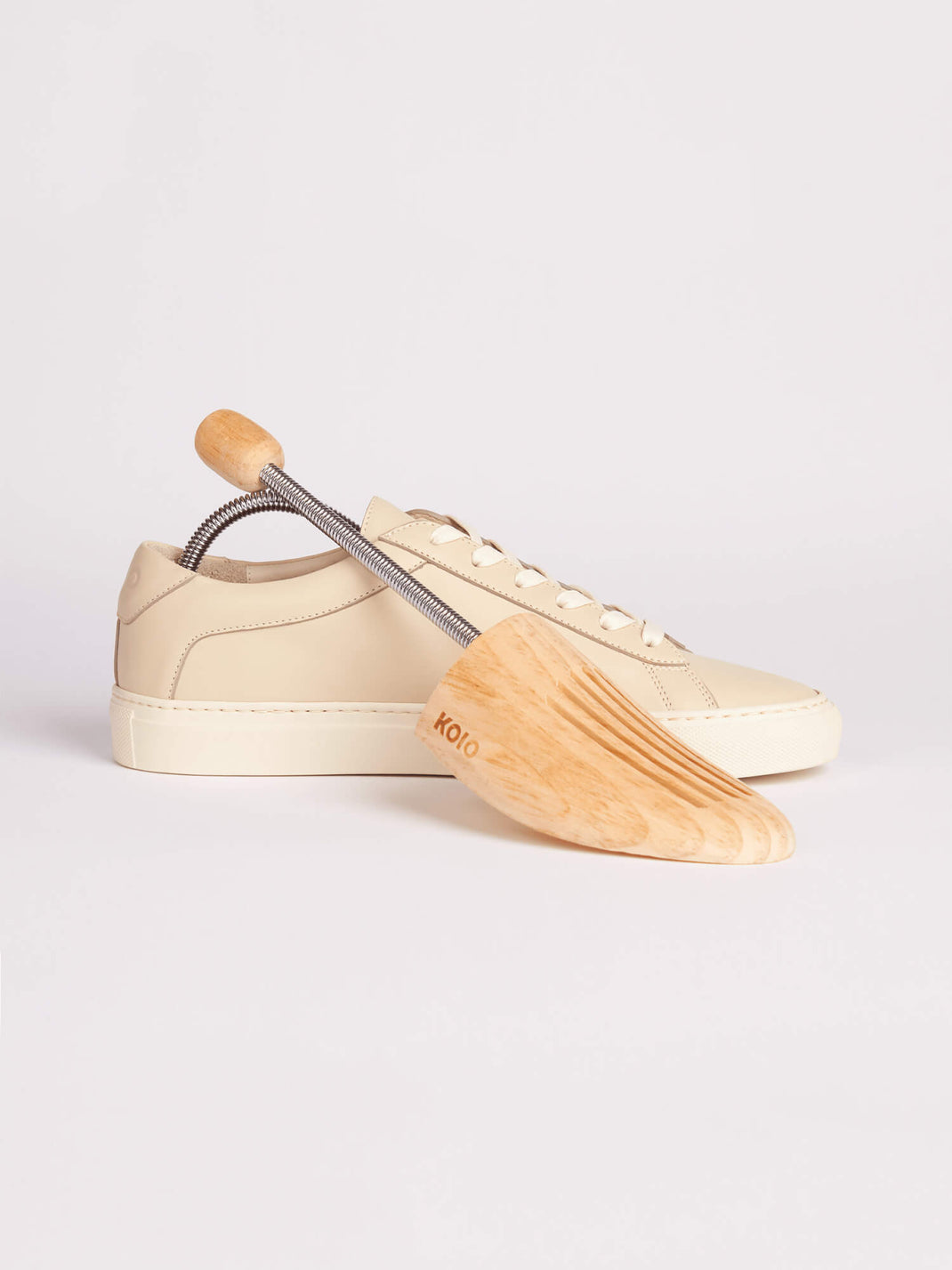 Side view of KOIO shoe tree showcasing its spring mechanism inside a cream low-top sneaker against a light background