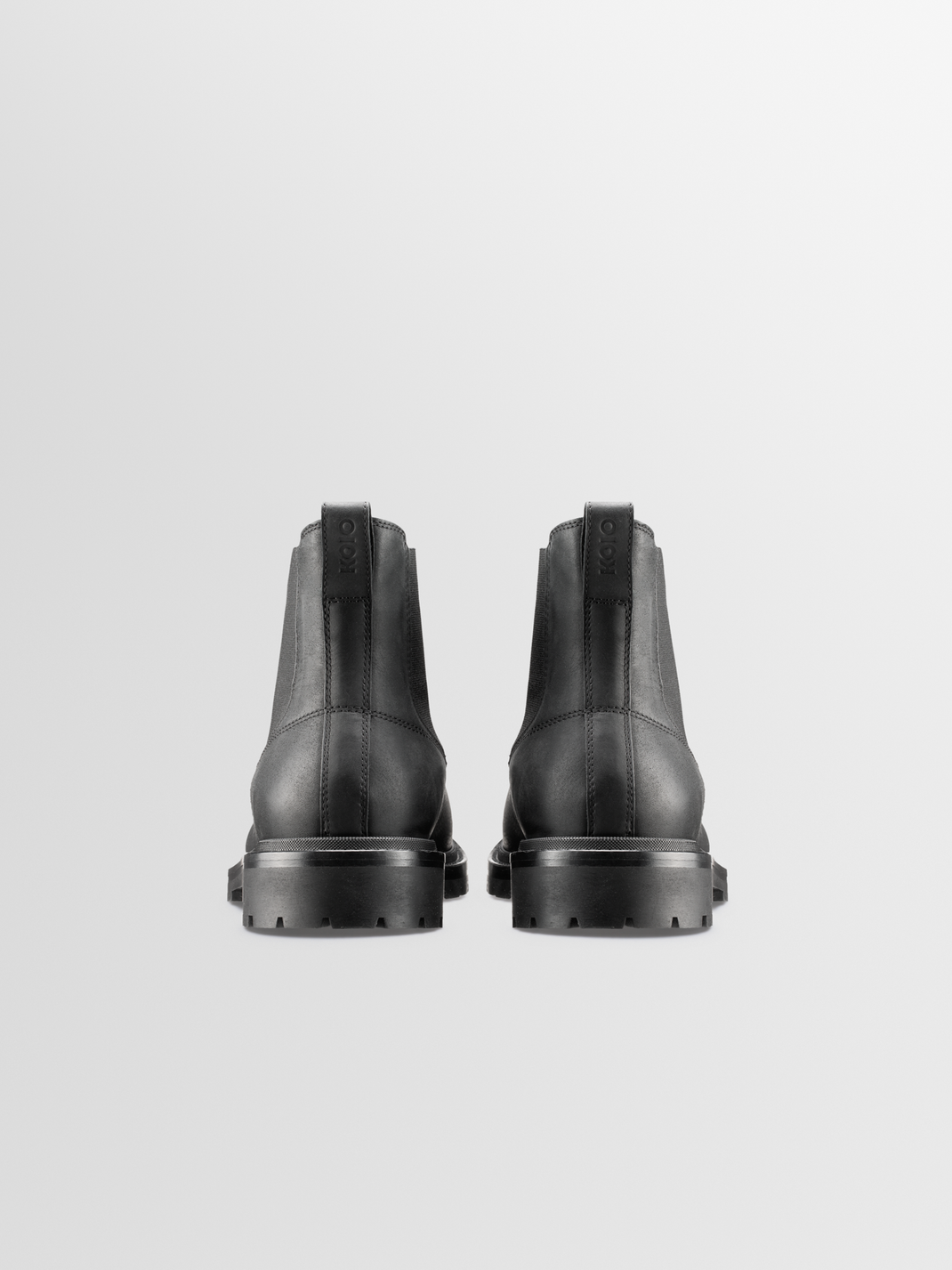 Rear view of black leather Chelsea boots showcasing pull tabs and lug soles with a light gray background